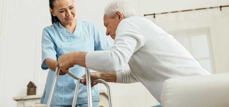 Person in scrubs helping elderly person with a walker.
