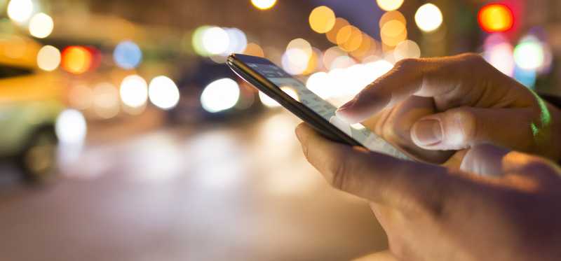 A hand holds a cell phone against a background of lights.