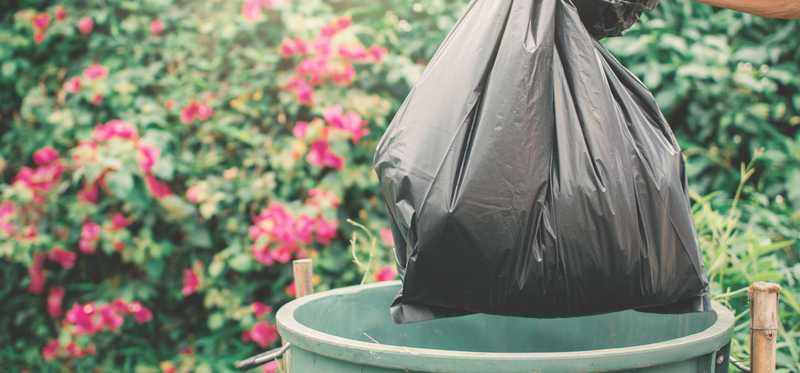 A garbage bag being thrown in the trash can with red flowers in the background.