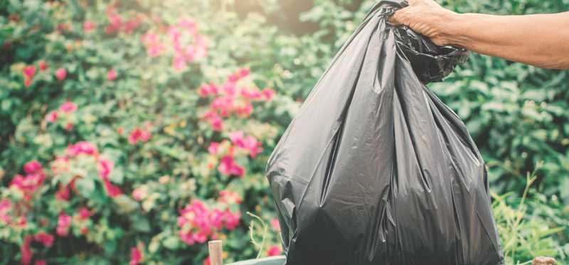 A garbage bag being thrown in the trash can with red flowers in the background