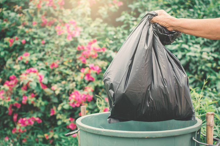 A garbage bag being thrown in the trash can with red flowers in the background