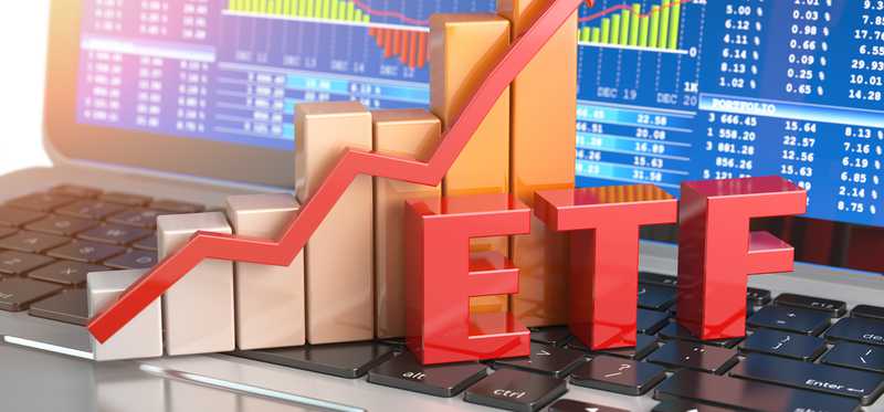 The abbreviation ETF with a bar chart and rising arrow behind it and all sitting on an open laptop with stock charts on the screen.