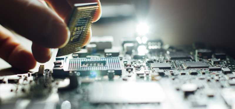 A hand holding a microchip above a circuit board.