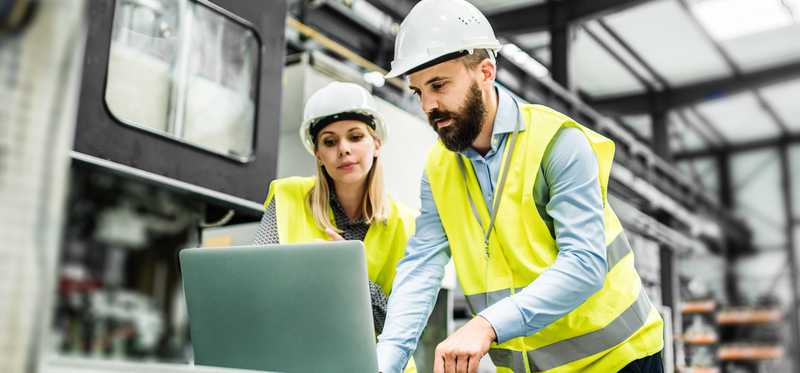 Two people in yellow vests and hard hats working in a factory setting.