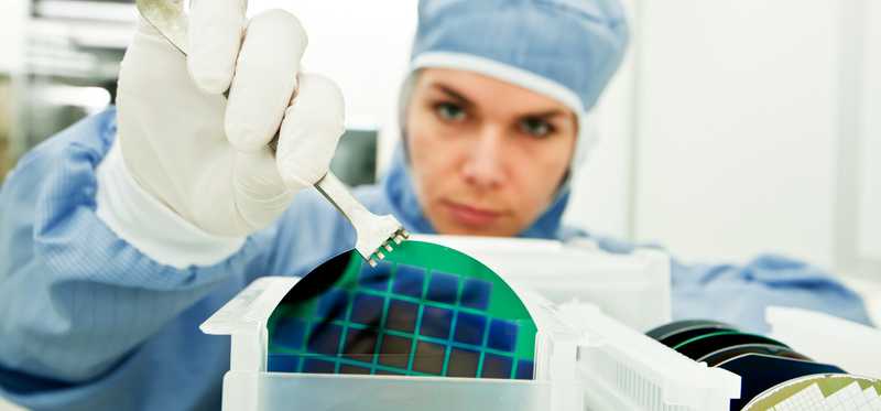 A woman in a dust free room using a tool to hold up a silicon wafer.