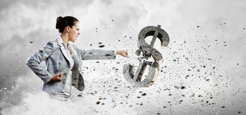 A woman in business attire punching a dollar sign that crumbles under her blow.