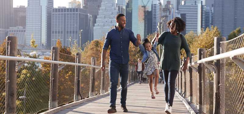 Two people swing a child between them as they all walk on a city footbridge.