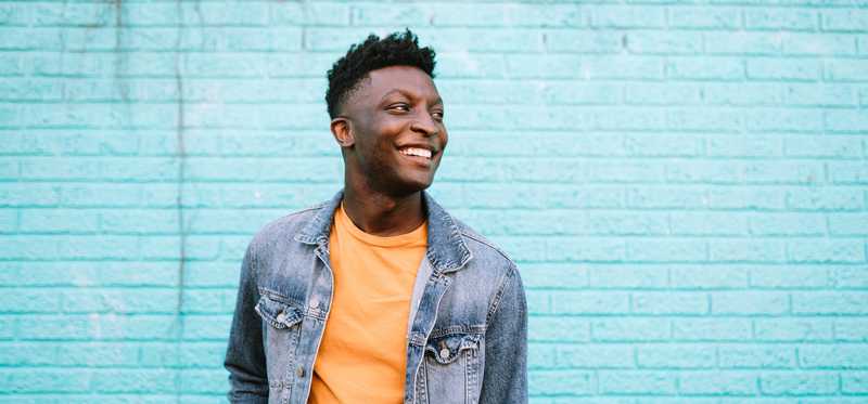 A smiling person standing in front of a turquoise brick wall.