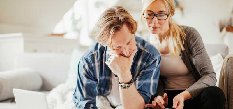 Two people using calculator and laptop while sitting on couch at home.