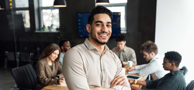 Professionally dressed individual smiles while leaning against conference room table, with other professionals seated behind.