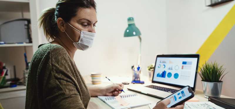 Masked woman sits at desk with open laptop while writing and looking at phone.