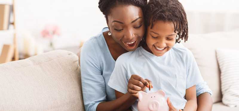 Smiling adult with child in lap helps child drop coins into a piggy bank.
