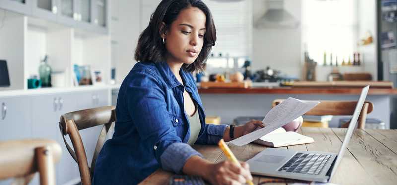 Person focused while taking notes and looking at paperwork in front of a laptop.