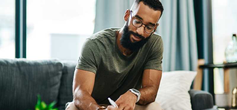 Person sitting on couch and writing on paperwork on coffee table.