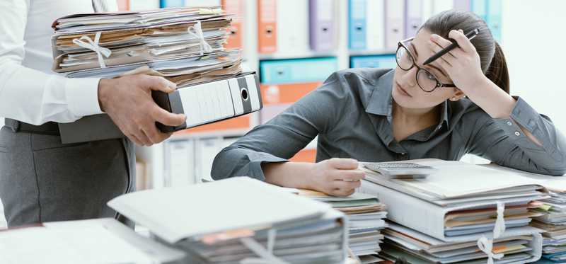 A frustrated woman with a desk full of paperwork and a coworker bringing more