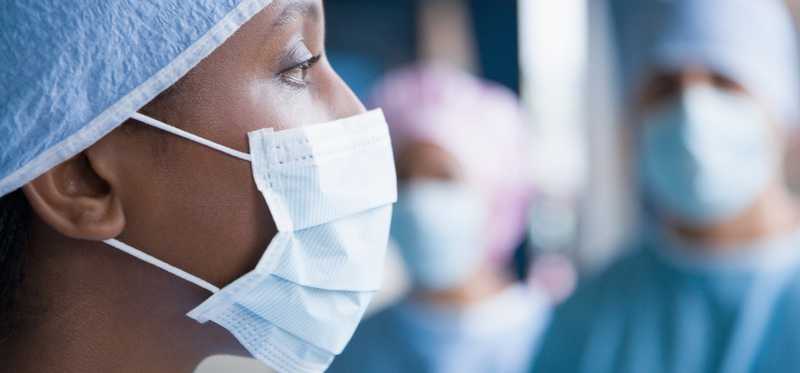 A person in a surgical mask and attire with similarly clad people nearby.