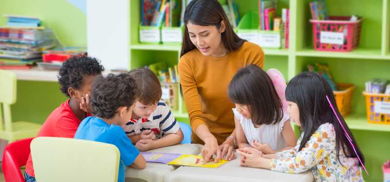 Adult sits in a brightly colored room with several children.