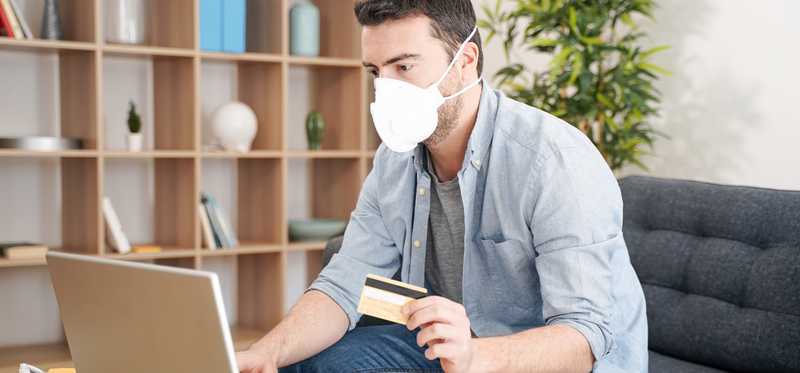 Person wearing mask holding credit card in front of laptop.
