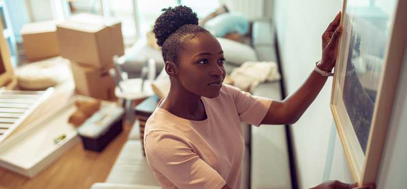 Smiling person hangs wall art in home with unpacked boxes.