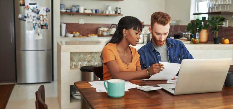 Two people looking at paperwork and open laptop in kitchen.