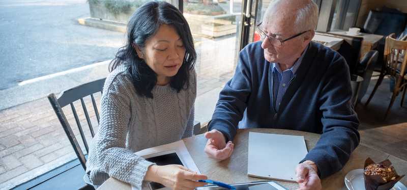 Two people reviewing paperwork and tablet at table by window.