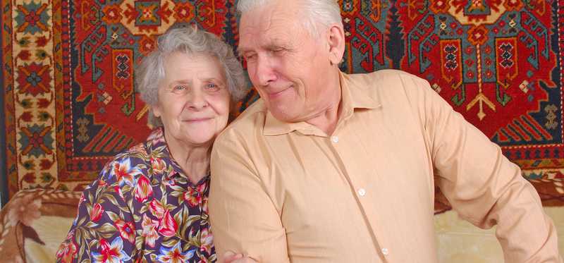 Elderly couple sit side by side smiling on a couch.