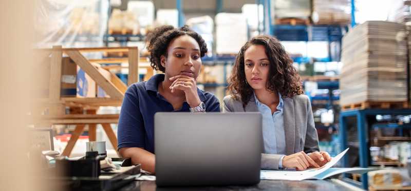 Two people working at a laptop on a table in a warehouse.