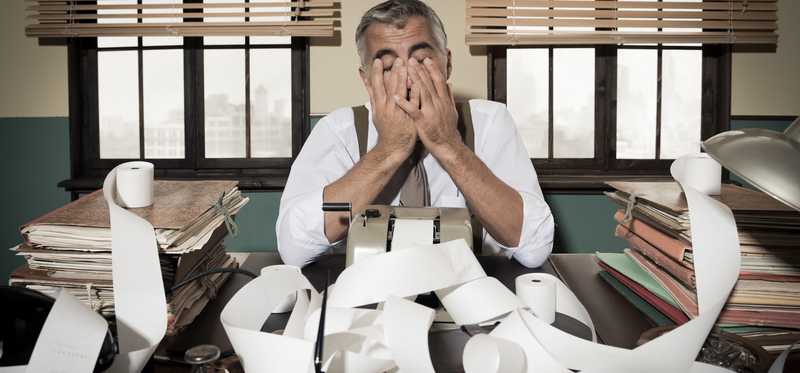 Person sits in old office building surrounded by folders and rolls of paper with face in hands.