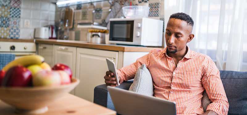 Person sitting on couch in kitchen while looking at phone and laptop.