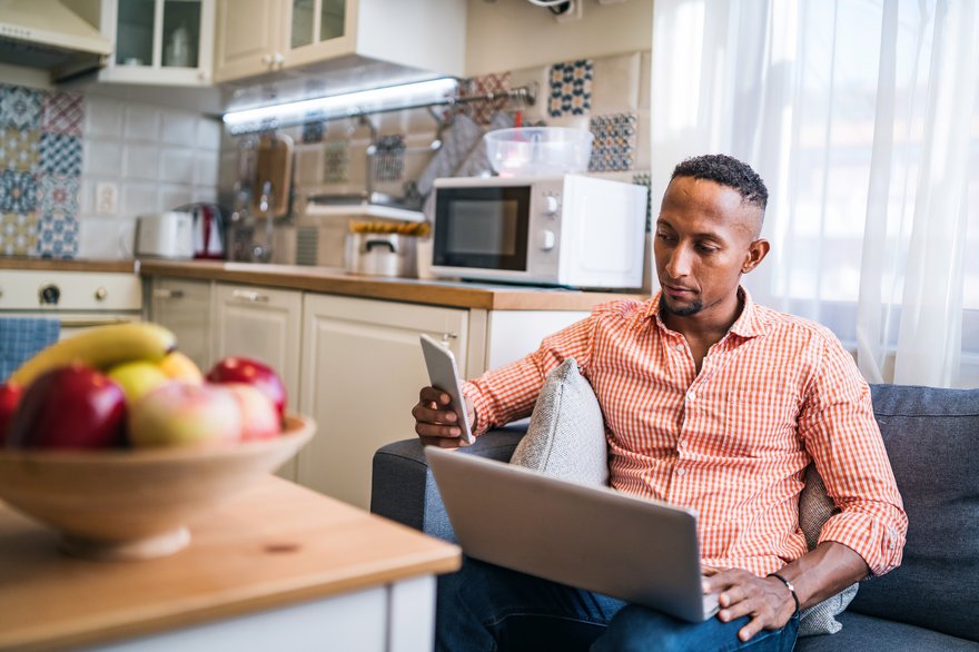 Person sitting on couch in kitchen while looking at phone and laptop.
