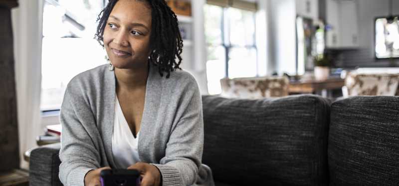 Adult smiles thoughtfully while sitting on couch at home with smartphone.