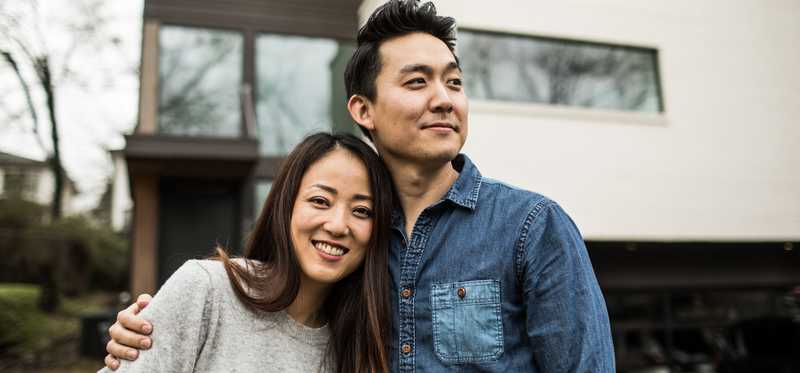 Couple stands in front of home smiling, while arm in arm.