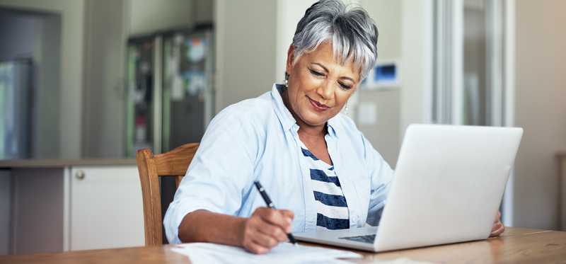 Smiling person takes notes in front of laptop at home.