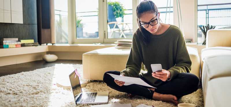 Person sitting with legs crossed on the floor is looking at phone and paperwork.