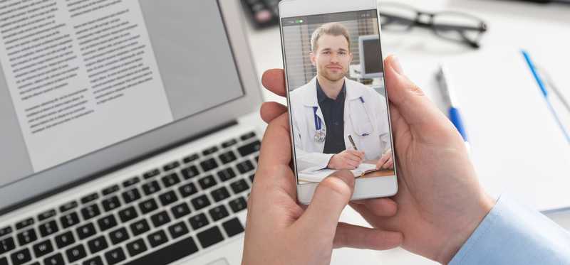 Person with laptop in front of them views doctor on smartphone screen.
