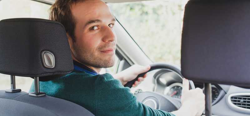 We see a young man in a car's driver's seat, turning around to look at someone in the back seat.