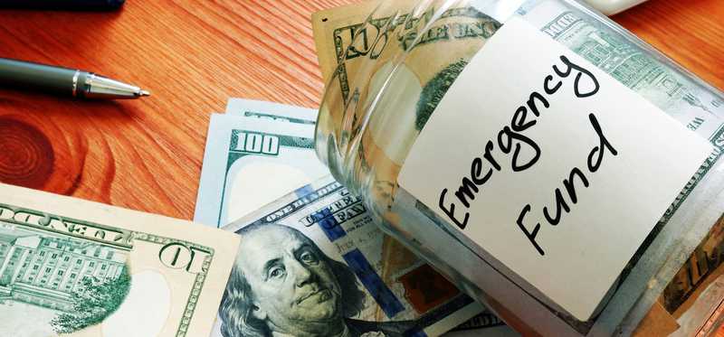 A desk with a calculator and a glass jar that says Emergency Fund and is full of cash.