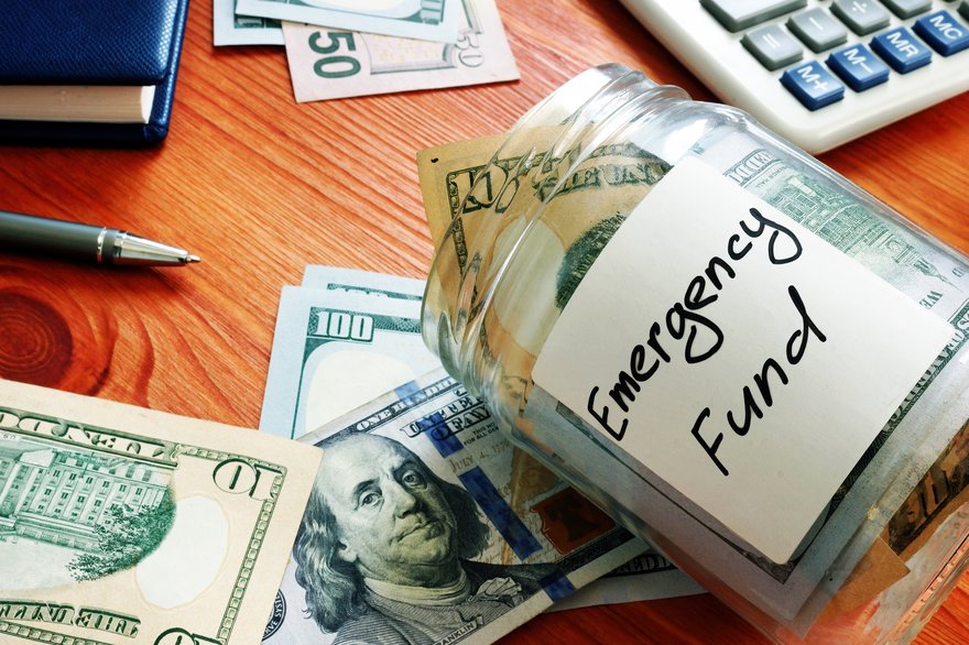 A desk with a calculator and a glass jar that says Emergency Fund and is full of cash.