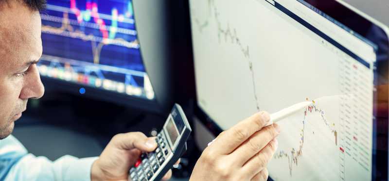 Person holds calculator and uses pencil to point to stock chart on computer screen.