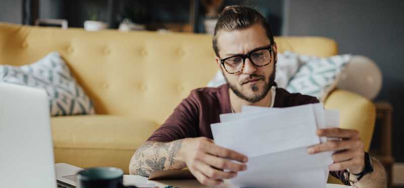 Person in glasses is looking at paperwork while seated on living room floor near couch.
