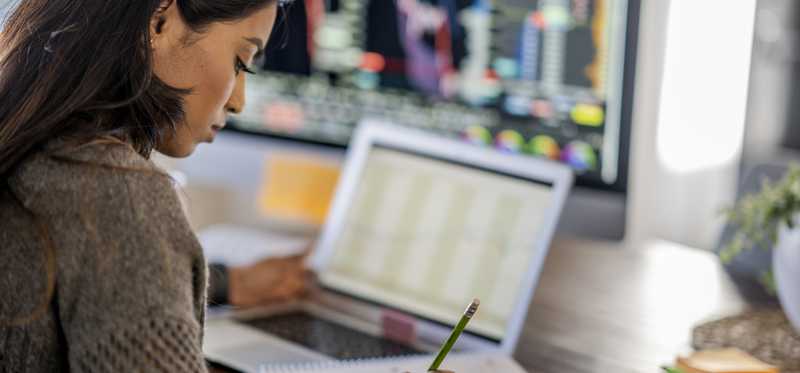 Person writing in notebook by computer screens.