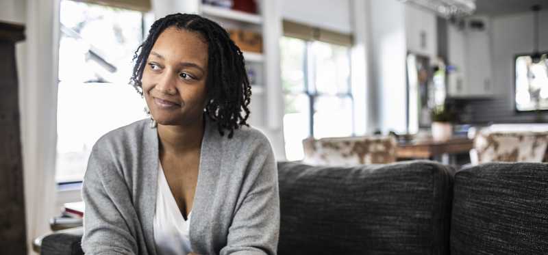 Person smiles thoughtfully while sitting on couch and holding smartphone.