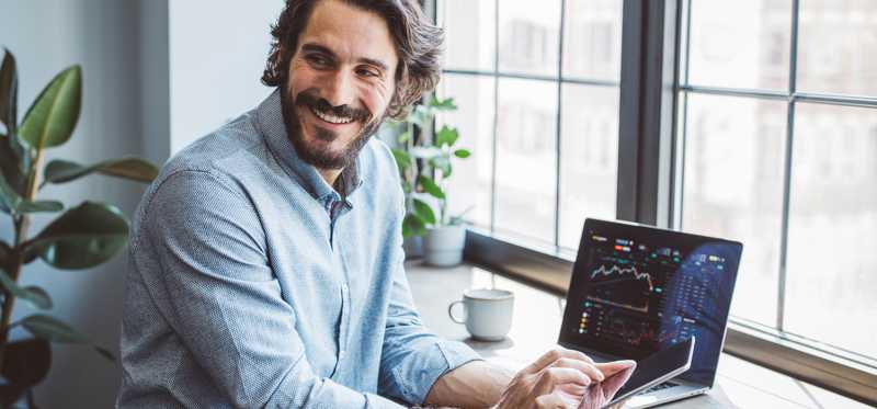 Person smiling while using phone and laptop by window.