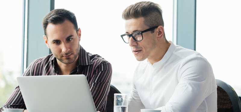 Two people looking at a laptop near large office windows.