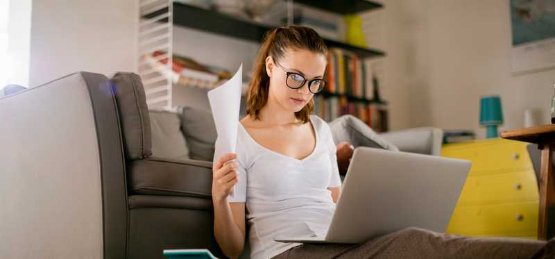 Person sitting on couch with laptop and paperwork.