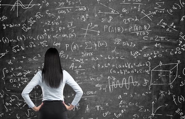 A young woman looks at a chalkboard full of numbers.