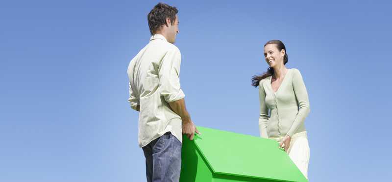 Two people lift a small green house in a sunny field.