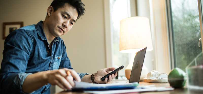 Person sits at window while working on laptop, paperwork and phone.