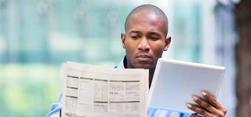 Person studies financial newspaper and tablet at desk.
