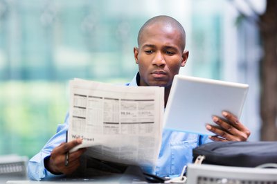 Person studies financial newspaper and tablet at desk.